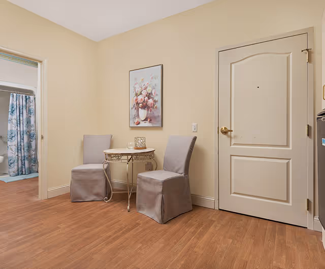 Small apartment entry area with two upholstered chairs and a round side table under a floral painting, a closed door to the right and a bathroom doorway visible to the left.