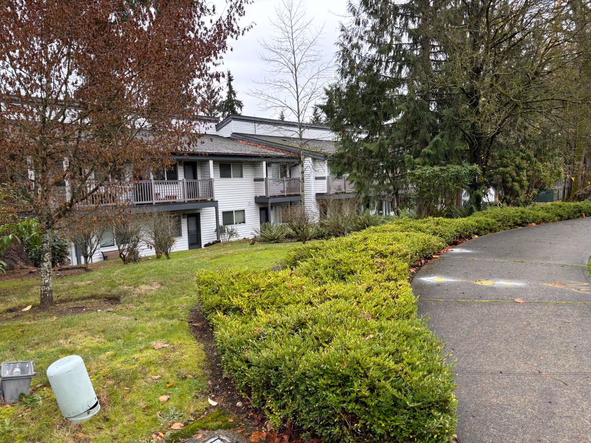 Two-story white residential building with balconies set behind shrubs, trees, and a curved driveway.