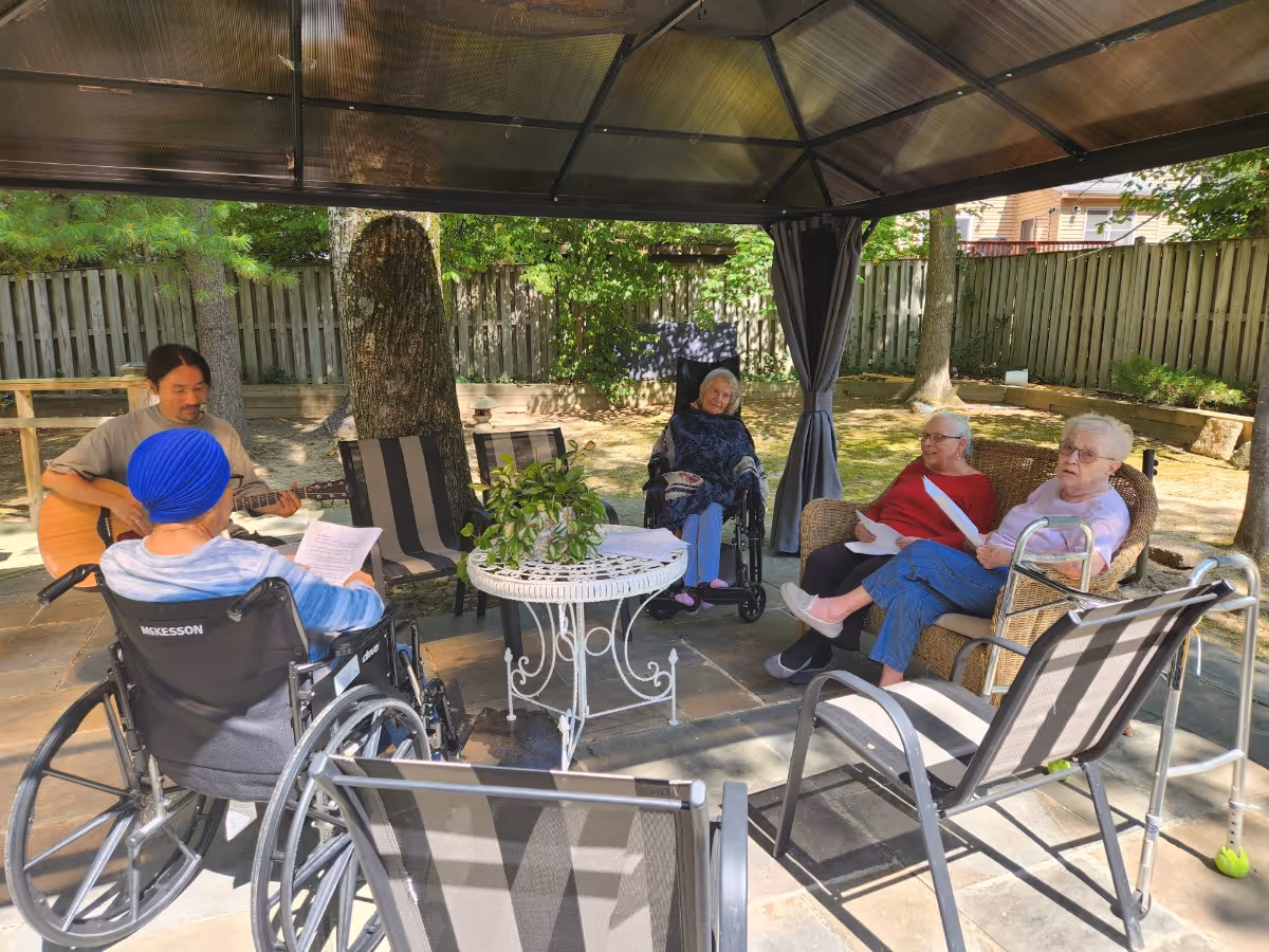 A group of elderly individuals sitting outdoors under a gazebo in a garden area. Two women are seated on wicker chairs, one woman is in a wheelchair, and another person in a wheelchair is holding a paper. A man is playing a guitar, and the group appears to be engaged in a musical or social activity. The area is surrounded by trees and a wooden fence.