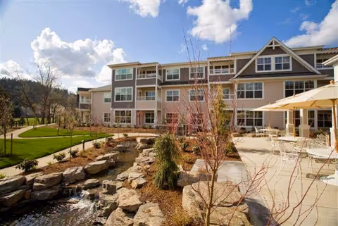 Outdoor view of The Stafford Retirement Community showing a landscaped garden with a small stream flowing over rocks, young trees, and a patio area with tables and umbrellas. The multi-story building with large windows is visible in the background under a partly cloudy sky.