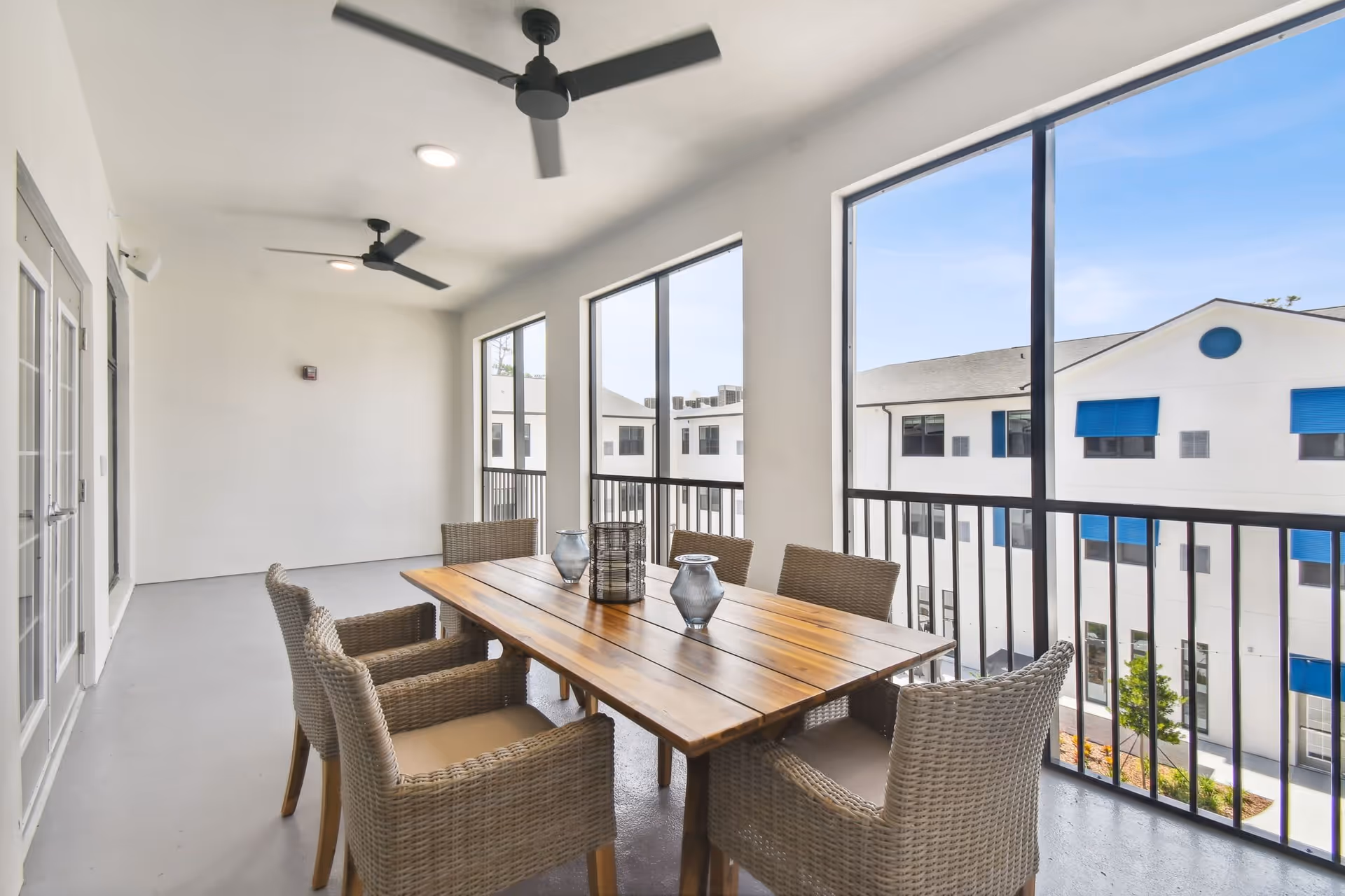 A covered outdoor balcony area with a wooden dining table surrounded by six wicker chairs. The balcony has large windows with black railings and ceiling fans above. Outside, white buildings with blue window awnings are visible under a clear sky.