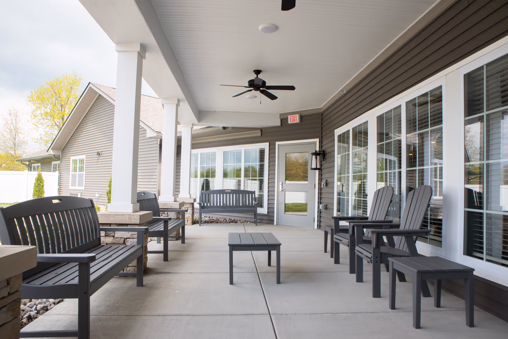 Covered outdoor patio area at Cooper Trail Senior Living with multiple dark gray benches, chairs, and small tables arranged on a concrete floor. The patio has white columns, a ceiling fan, and large windows with white frames along the building wall. A door with an exit sign is visible at the far end.