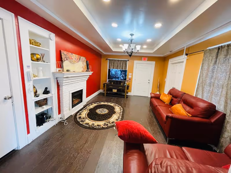 A cozy living room with red leather sofas adorned with orange and red pillows. The room features a white brick fireplace against a red accent wall, a decorative round rug on dark wooden flooring, and a TV on a stand in front of a window with patterned curtains. The walls are painted yellow and the ceiling has recessed lighting and a chandelier.