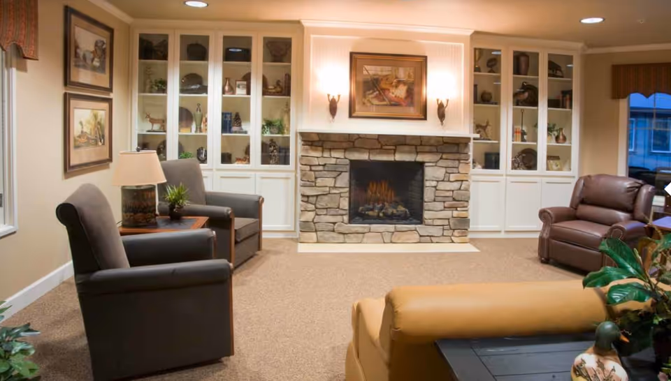 A cozy living room with a stone fireplace in the center, flanked by built-in white cabinets with glass doors displaying decorative items. The room has beige carpet and walls, two gray armchairs with a small wooden table and lamp between them on the left, a brown leather armchair on the right, and a tan sofa in the foreground. There are framed pictures on the wall and a window with a valance on the right side.