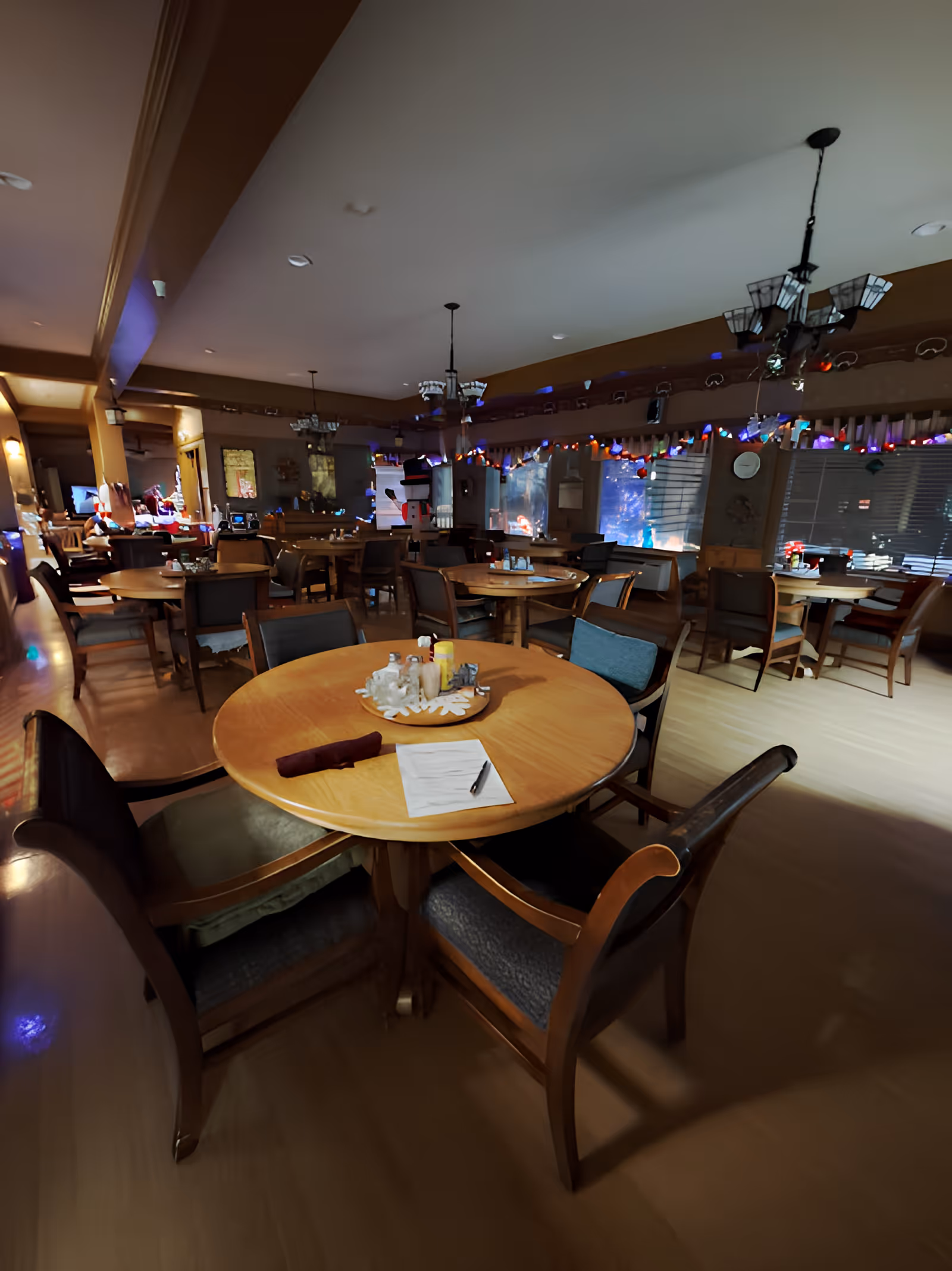 Interior view of a dining room with multiple round wooden tables and chairs arranged neatly. The room is decorated with string lights and holiday decorations, including a snowman figure near the windows. The lighting is warm and cozy, creating a welcoming atmosphere.