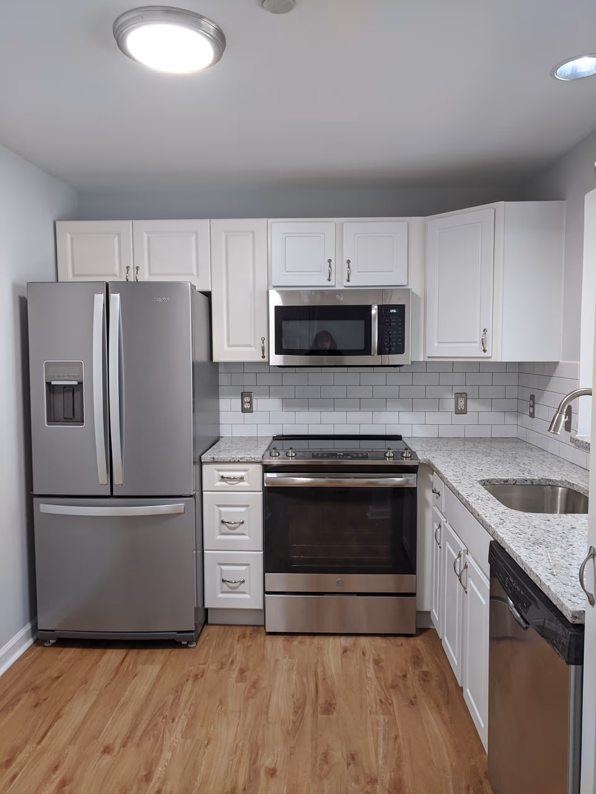 Modern kitchen with stainless steel refrigerator, oven, microwave, dishwasher, white cabinets, granite countertops, and wood flooring.
