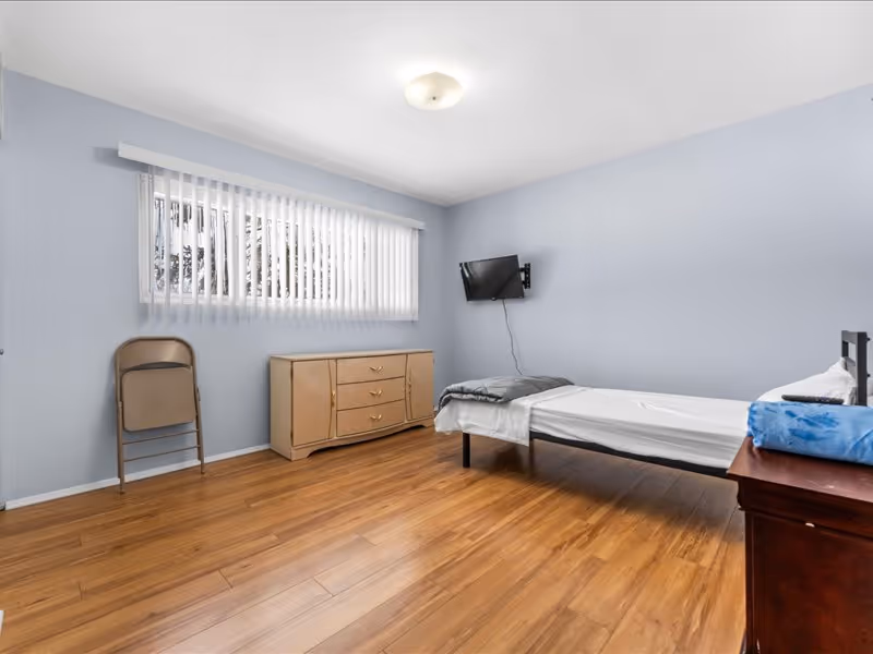 A simple bedroom with light blue walls and wooden flooring. The room contains a single bed with white sheets and a gray blanket, a small wooden nightstand with a blue towel on it, a beige dresser with three drawers, a folded beige chair leaning against the wall, and a wall-mounted flat-screen TV. A window with vertical blinds allows natural light into the room.