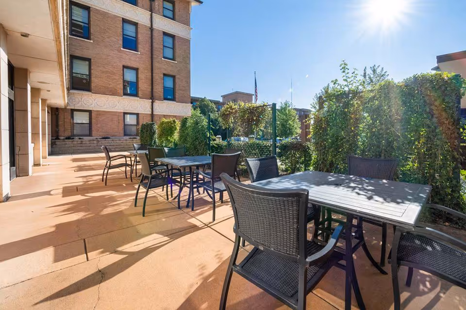 Outdoor patio area at EverTrue Laclede Groves with multiple tables and chairs arranged for seating. The patio is adjacent to a brick building and surrounded by greenery and bushes under a clear blue sky with sunlight.