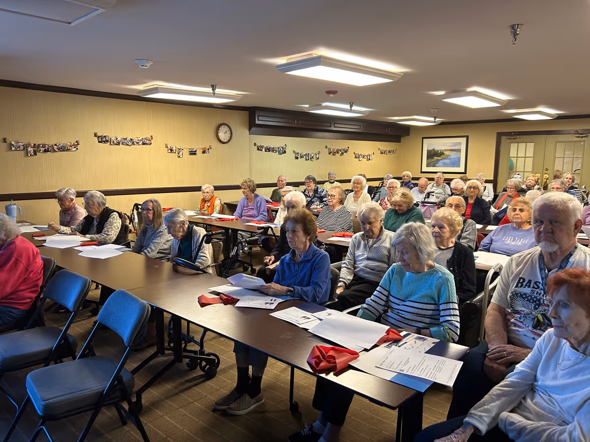A group of elderly people seated in a classroom-style setting at Atria Kinghaven, attentively looking at papers in front of them. The room has beige walls with photos hanging on strings, a clock, and a framed picture. The lighting is bright with ceiling lights.