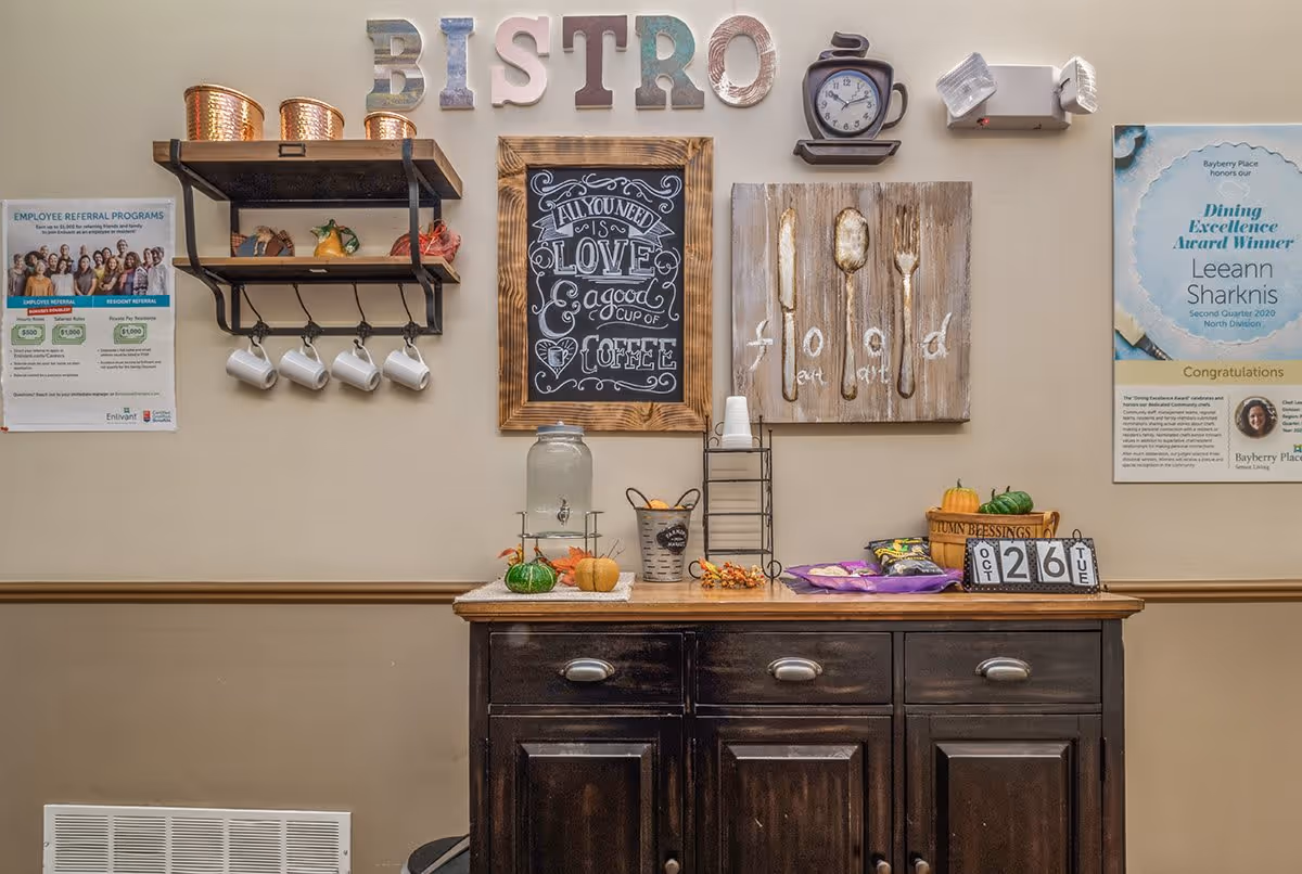 Bistro-style serving station with a wooden cabinet, chalkboard sign, mugs, snacks, and decorative wall art.