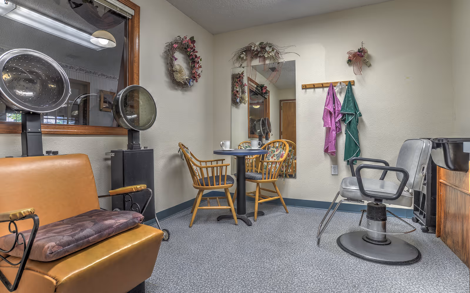 Interior salon room with hairdryers and styling chairs, a small table and chairs, mirror and hanging robes.