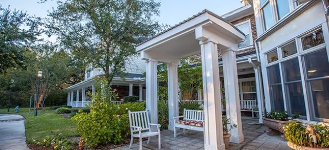 Front exterior courtyard with a covered entryway supported by white columns, benches, and landscaped paths beside a multi-story building.