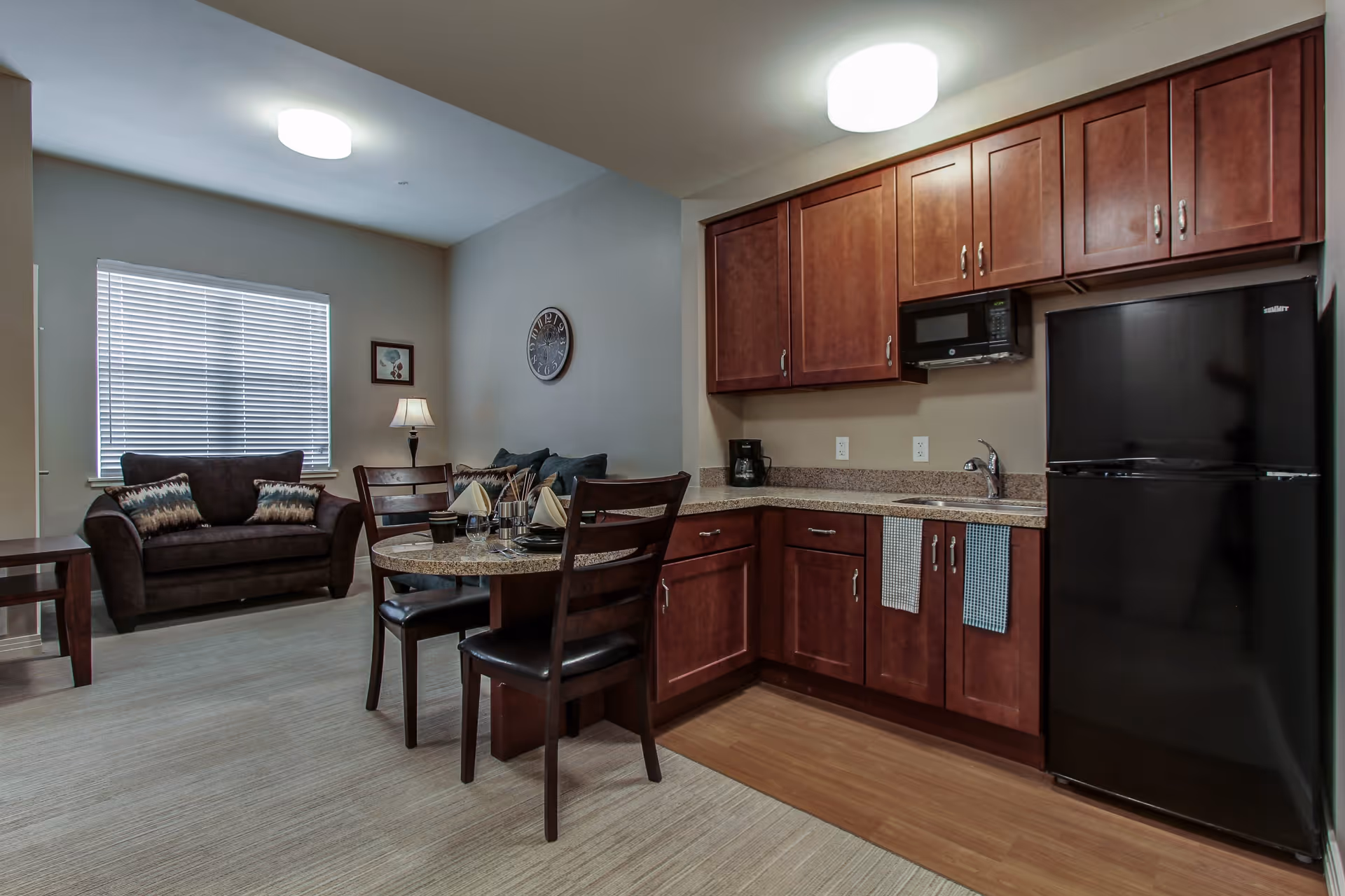 Interior view of a senior living facility apartment featuring a small kitchen area with wooden cabinets, a black refrigerator, microwave, and coffee maker. Adjacent to the kitchen is a round dining table set with two chairs, napkins, and tableware. In the background, there is a living area with a dark brown loveseat, a side table with a lamp, a wall clock, and a window with blinds.