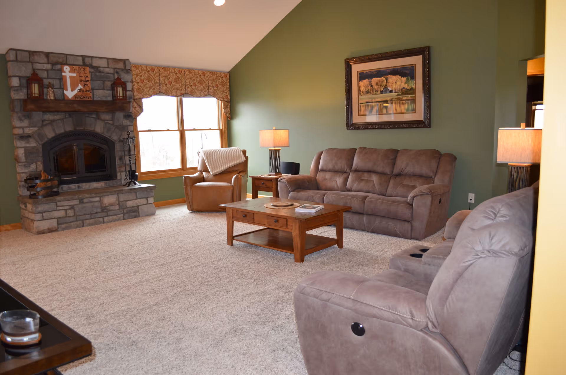 Living room with plush brown sofas and an armchair around a wooden coffee table, a stone fireplace, and a large window.