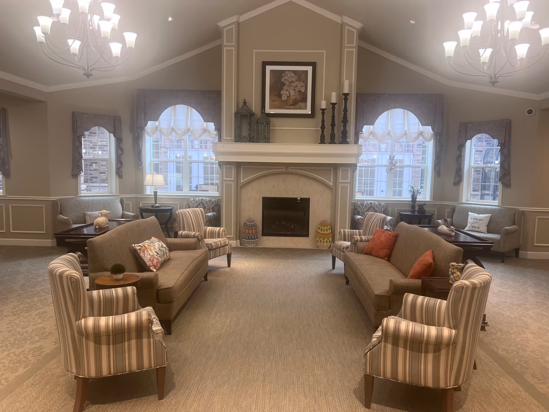 A cozy living room area in a senior living facility featuring two beige sofas with decorative pillows, four striped armchairs, a fireplace with decorative candles and framed artwork above it, and large windows with valances allowing natural light to fill the room. The room is carpeted and has soft lighting from chandeliers.