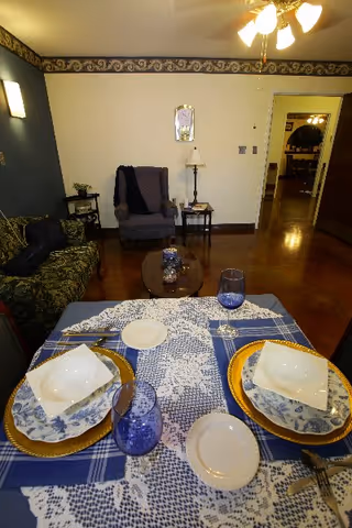 A cozy dining area set for two with blue and white patterned tablecloth and plates, blue glasses, and gold chargers. In the background, there is a green patterned couch, a gray armchair with a dark throw, a small side table with a lamp, and a hallway leading to another room.