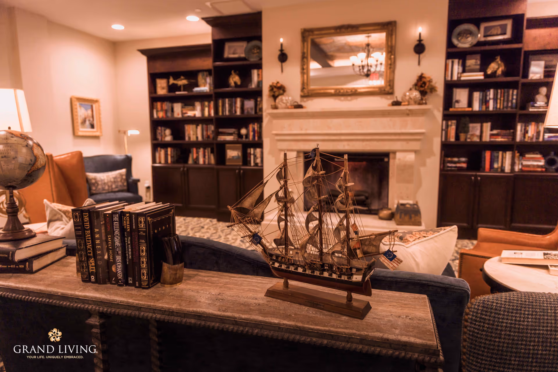 A cozy living room area with a model ship on a wooden table in the foreground, books and a globe also on the table. In the background, there are built-in dark wood bookshelves filled with books and decorative items, a fireplace with a large ornate mirror above it, and comfortable seating including a blue armchair and a brown leather chair.