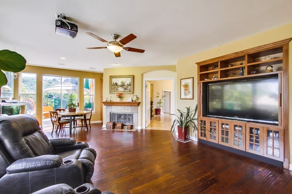 A spacious living room with wooden flooring, a large flat-screen TV in a wooden entertainment center, a black leather recliner chair, a ceiling fan with lights, and a fireplace with decorative items on the mantel. There is a dining table with chairs near large windows that let in natural light and offer a view of an outdoor patio with an umbrella and greenery.