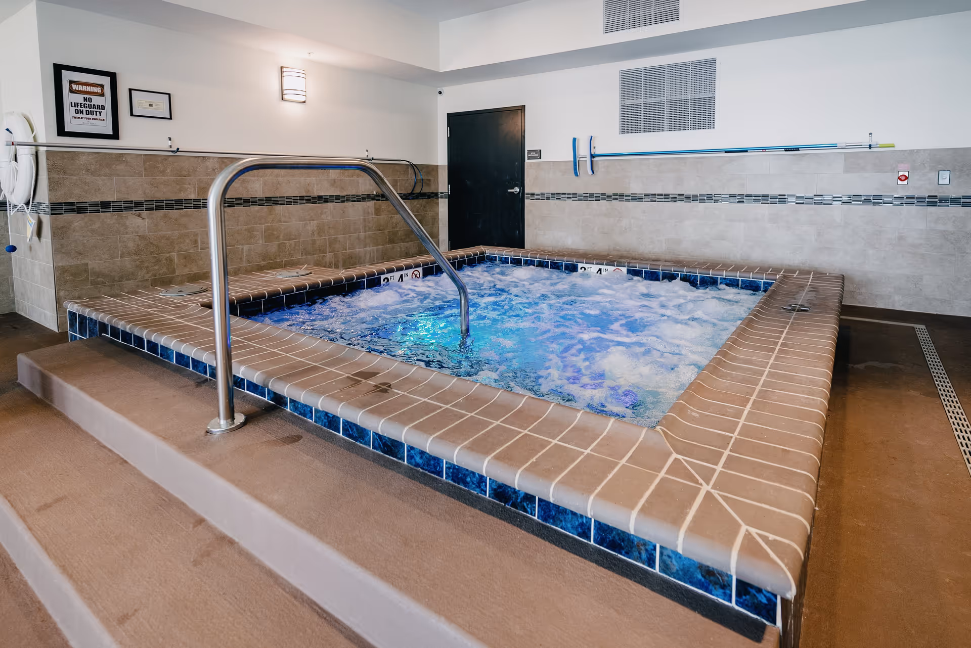 An indoor bubbling hot tub with tiled surround, metal handrail and steps in a community facility.