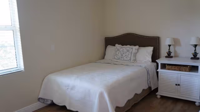 A simple bedroom with a single bed covered in a white quilt and a decorative pillow. Next to the bed is a white nightstand with two matching lamps and a wicker basket on the shelf. A window with blinds is on the left wall, allowing natural light into the room.