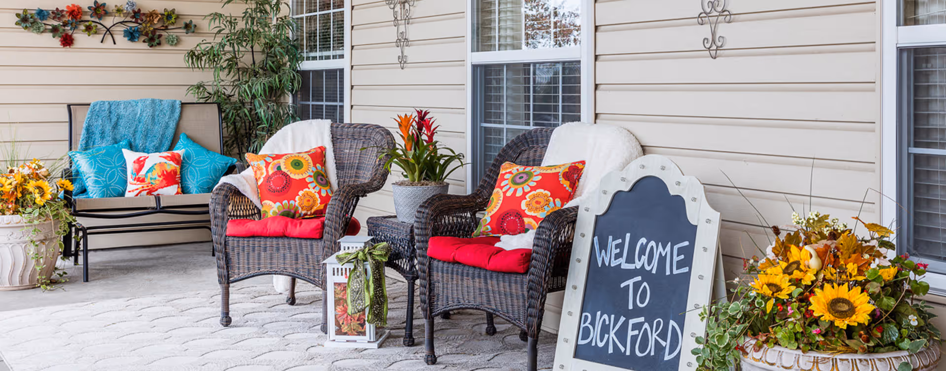 Outdoor patio area with two wicker chairs featuring red cushions and colorful floral pillows, a small table with a potted plant, a bench with blue and red pillows, decorative wall hangings, and a chalkboard sign that reads 'WELCOME TO BICKFORD' surrounded by flower pots with yellow and orange flowers.