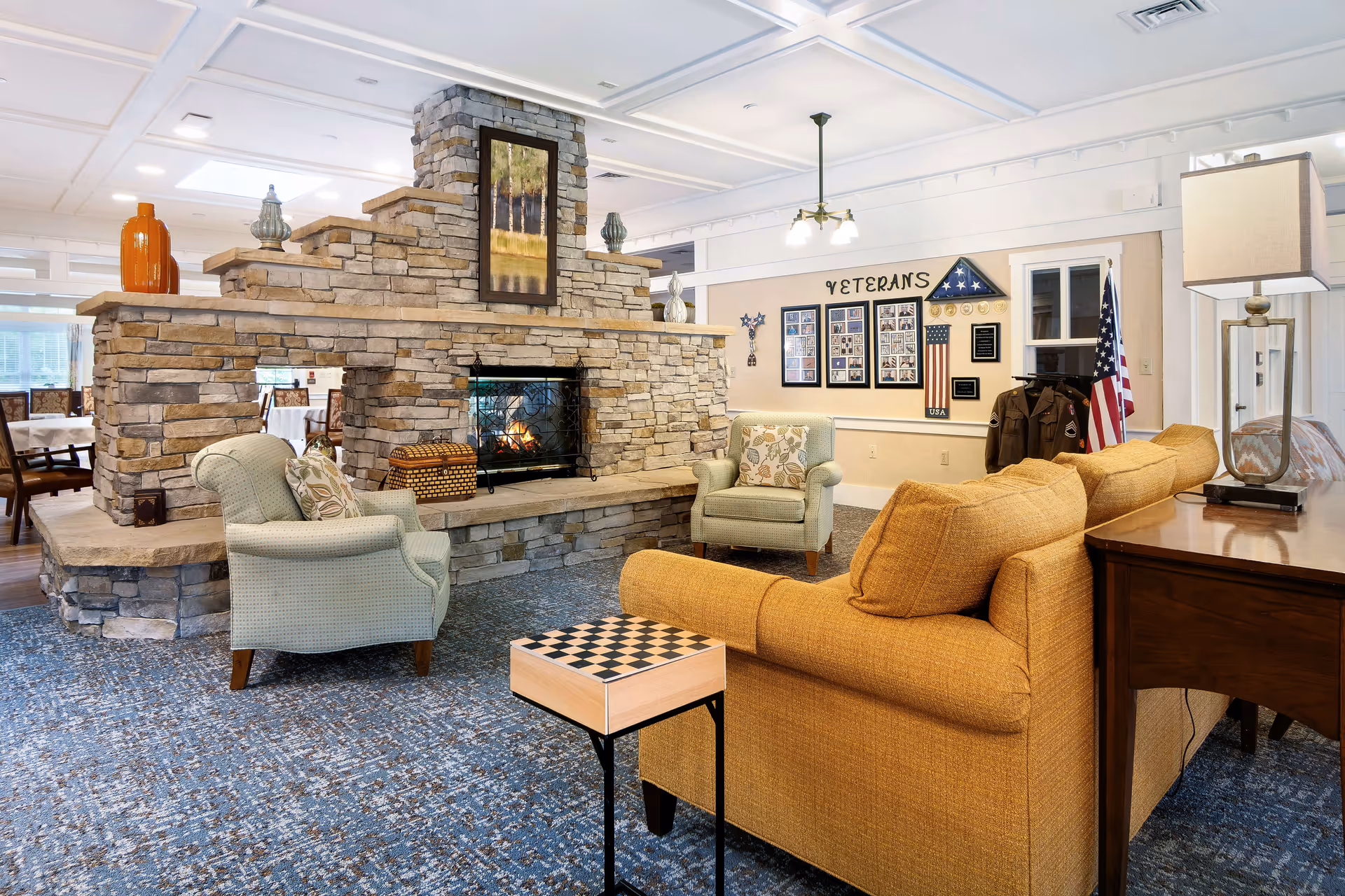 Cozy communal living room with a stone double-sided fireplace, armchairs and a mustard sofa, a veterans display and an American flag.