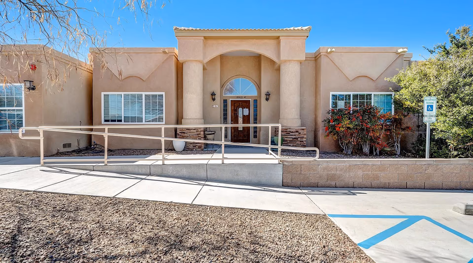 Front exterior view of Cottonwood Home, a single-story building with beige stucco walls, two large columns flanking the entrance, a wooden door with glass panels, and a wheelchair accessible ramp leading to the entrance. There are windows with white shutters on either side of the door, some bushes and trees, and a reserved parking space for handicapped near the entrance.