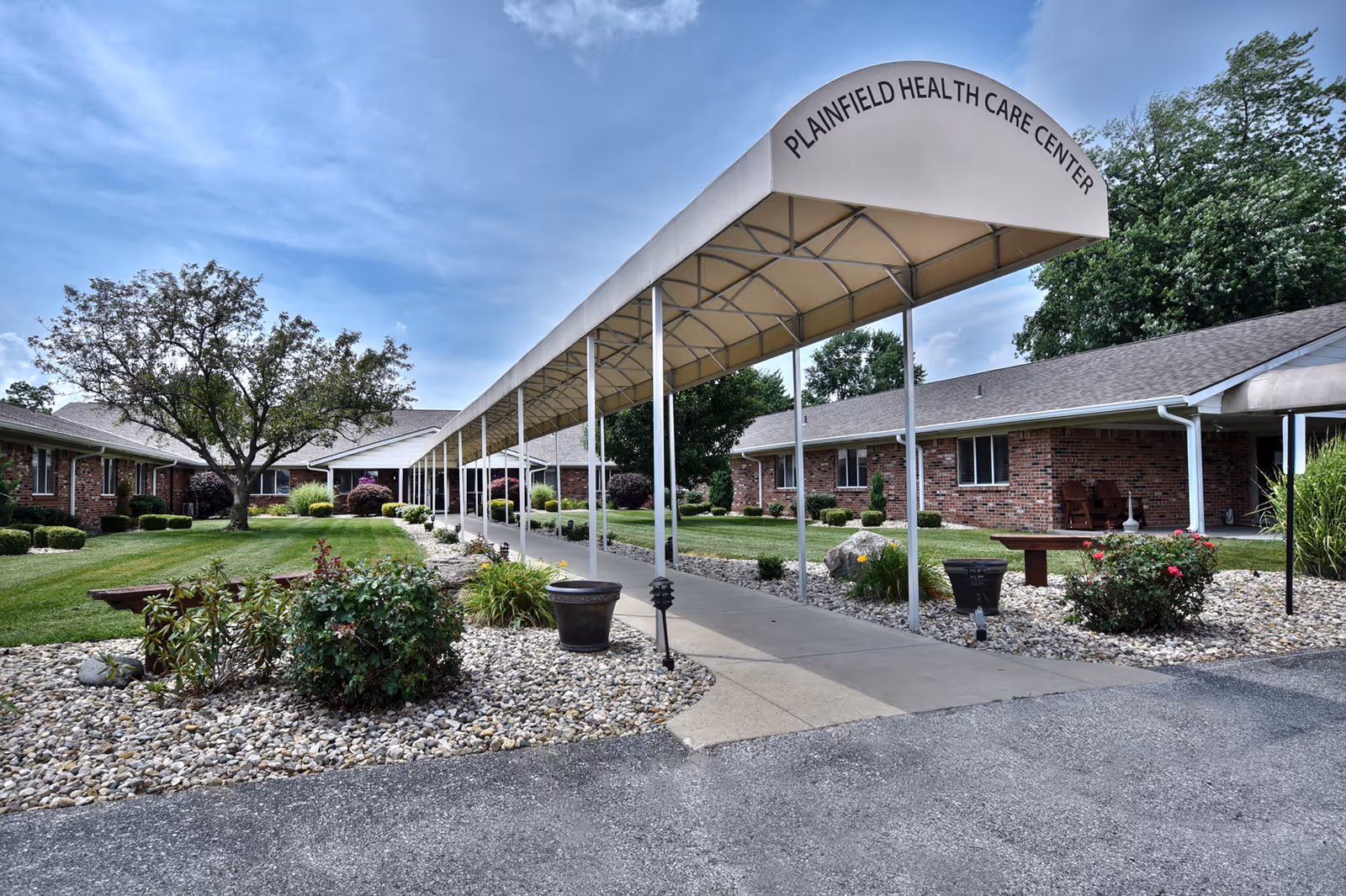 Exterior view of Plainfield Health Care Center showing a covered walkway leading to the entrance, surrounded by landscaped gardens with bushes, flowers, and trees under a partly cloudy sky.