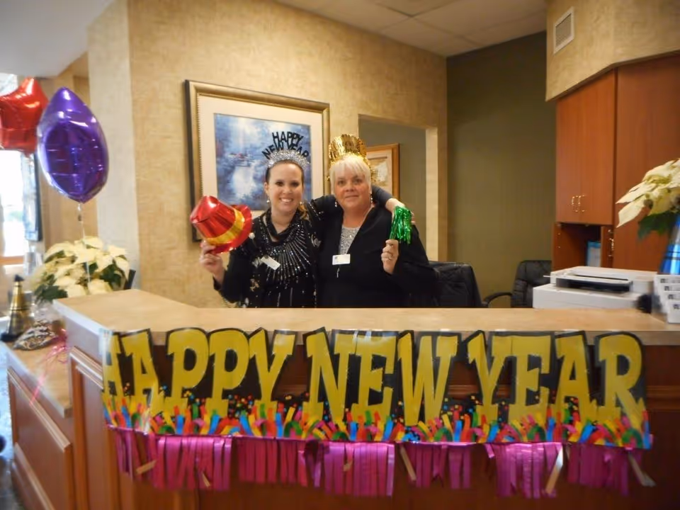 Two women standing behind a reception desk decorated with a colorful 'Happy New Year' banner. One woman is holding a red and yellow party hat, and the other is holding a green party horn. Both are wearing festive New Year headbands. Balloons and poinsettia plants are visible in the background.