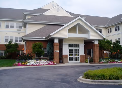 Front exterior view of a multi-story senior living facility with a covered entrance, brick and beige siding, surrounded by landscaped flower beds and greenery.