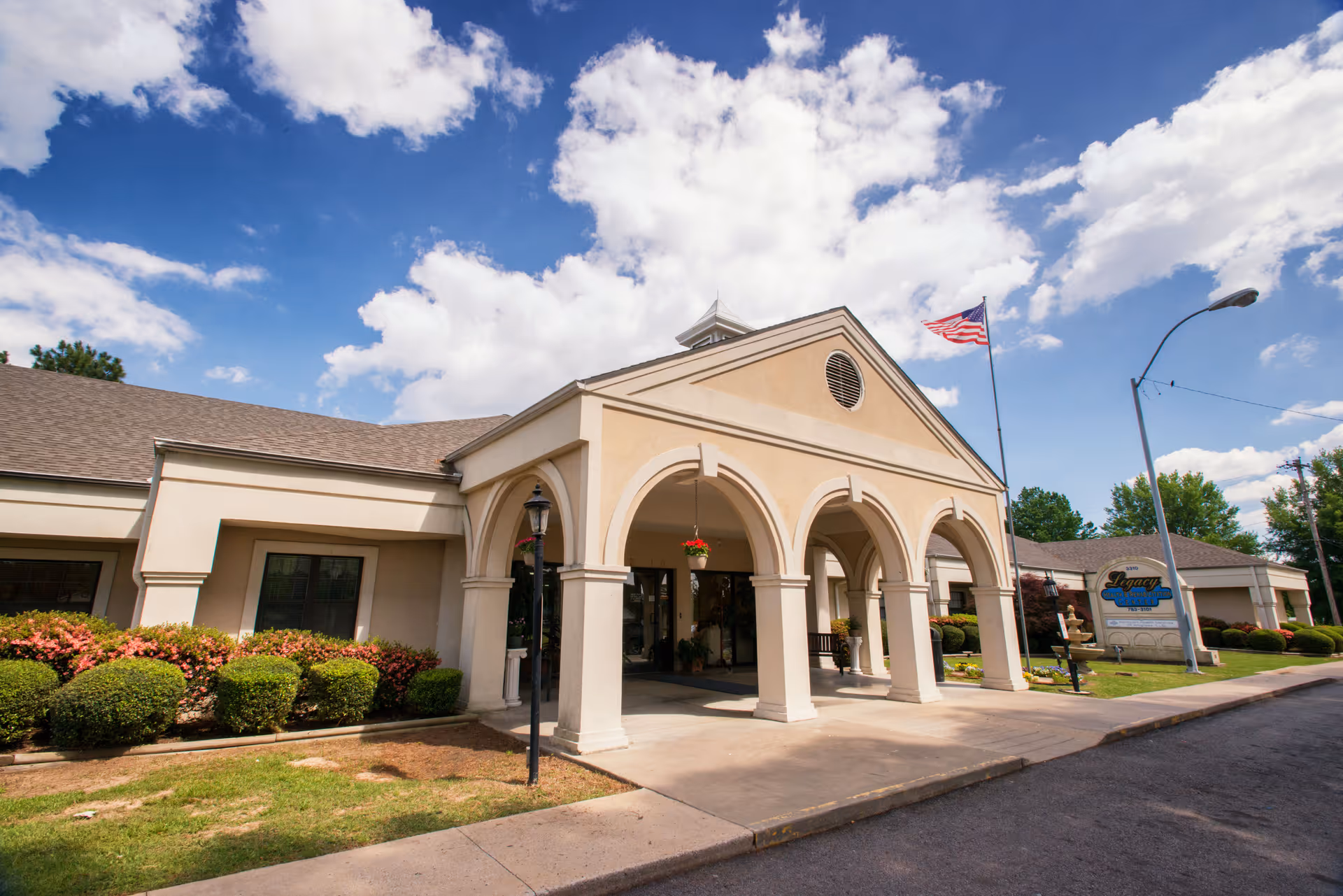 Exterior view of Legacy Health and Rehabilitation Center building with beige walls, arched entrance, and an American flag on a flagpole. The sky is partly cloudy and there are bushes and a lamp post near the entrance.