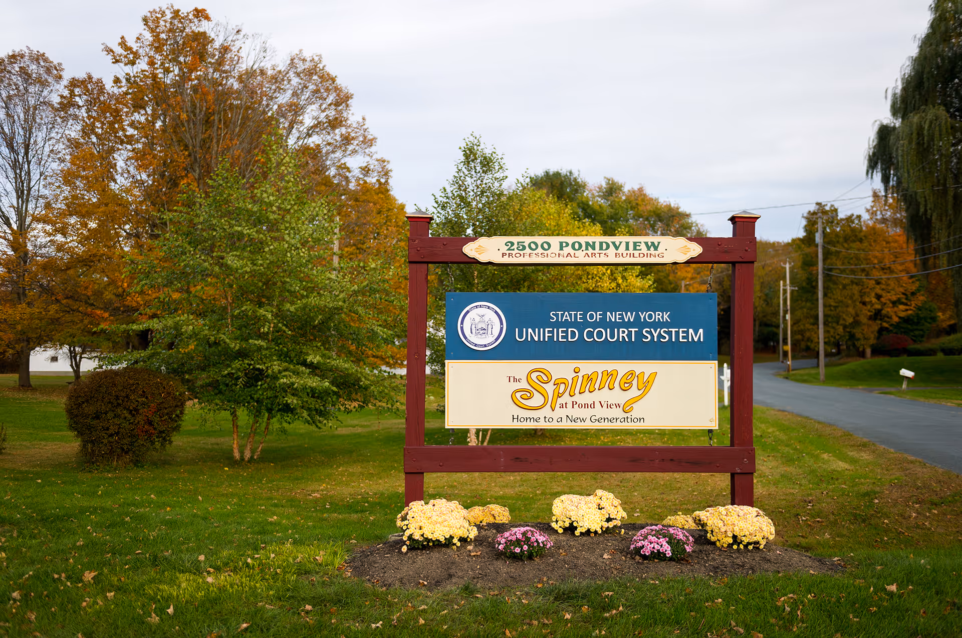 Wooden roadside sign for The Spinney at Pond View on a grassy lawn with trees and flowers.