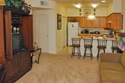 Interior view of a senior apartment showing a living room area with a wooden entertainment center holding a TV, a brown sofa, and a kitchen with wooden cabinets, a white refrigerator, microwave, and a counter with four bar stools.