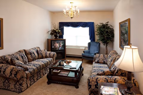 A cozy living room with two patterned sofas, a blue armchair, a wooden coffee table with magazines and decorative items, a television on a stand, a floor lamp, a chandelier, a large window with blue curtains, and a potted plant in the corner.