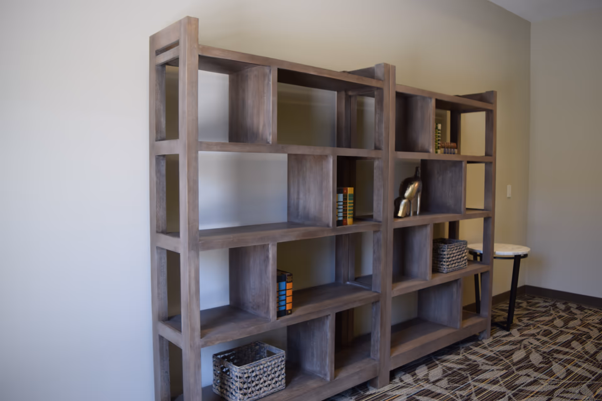 A wooden open bookshelf with multiple compartments, some holding books, decorative items, and baskets, placed against a beige wall with a patterned carpeted floor and a small round side table nearby.