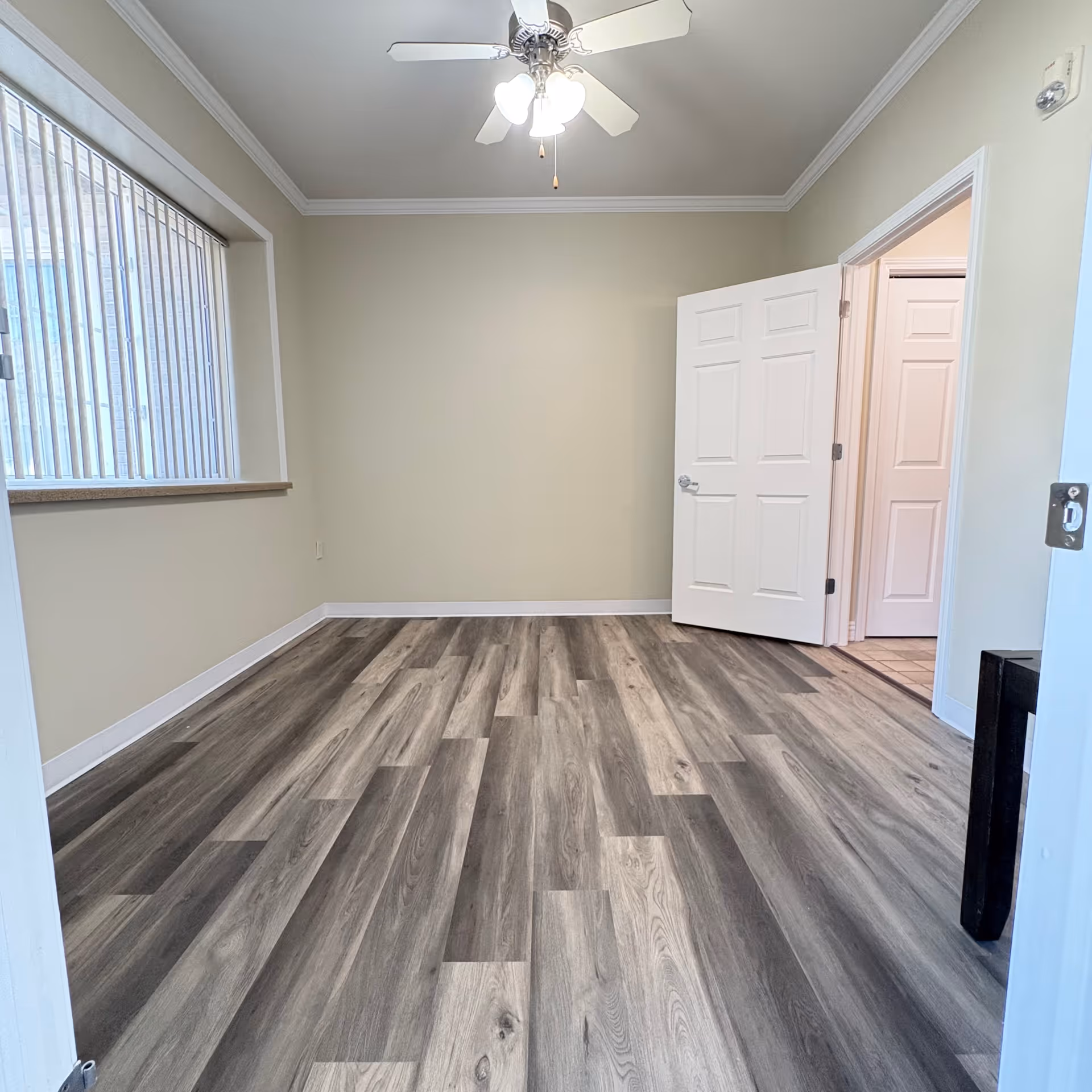 Empty room with light gray walls, wood-patterned vinyl flooring, a ceiling fan with lights, a large window with vertical blinds, and an open white door leading to another room.