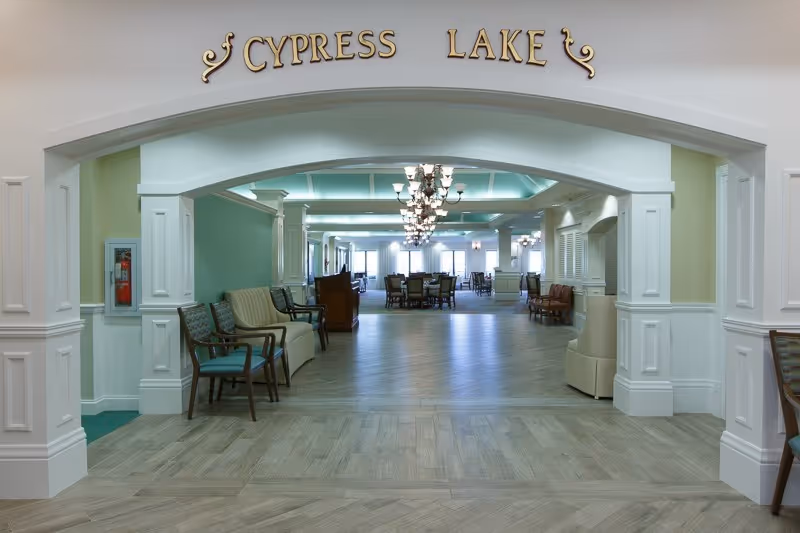View through a large white archway labeled 'CYPRESS LAKE' into a spacious, well-lit common area with multiple seating arrangements including chairs and tables, chandeliers hanging from the ceiling, and large windows in the background.