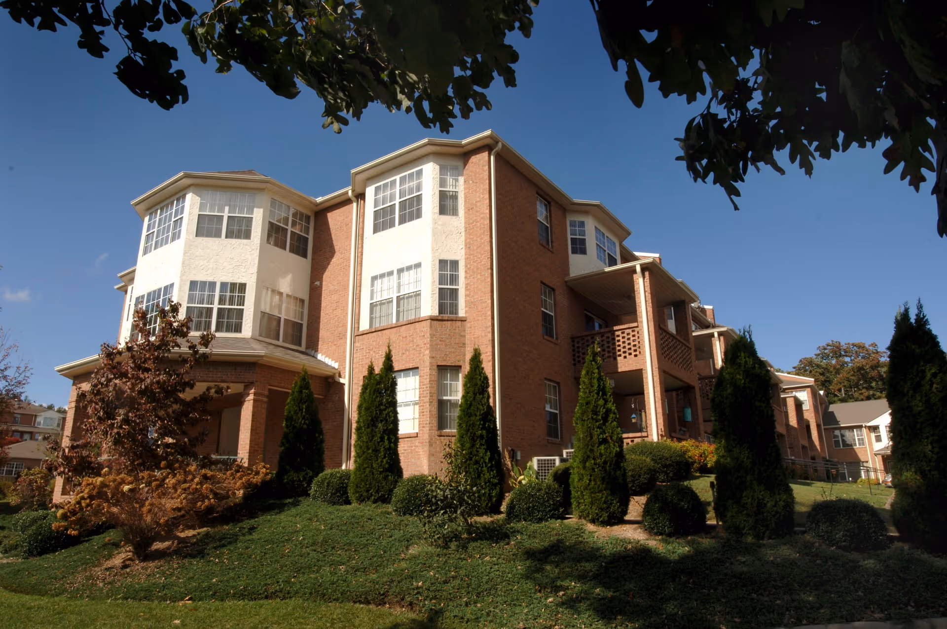 Exterior view of a multi-story brick and stucco building with large windows, surrounded by well-maintained landscaping including bushes and tall evergreen trees under a clear blue sky.