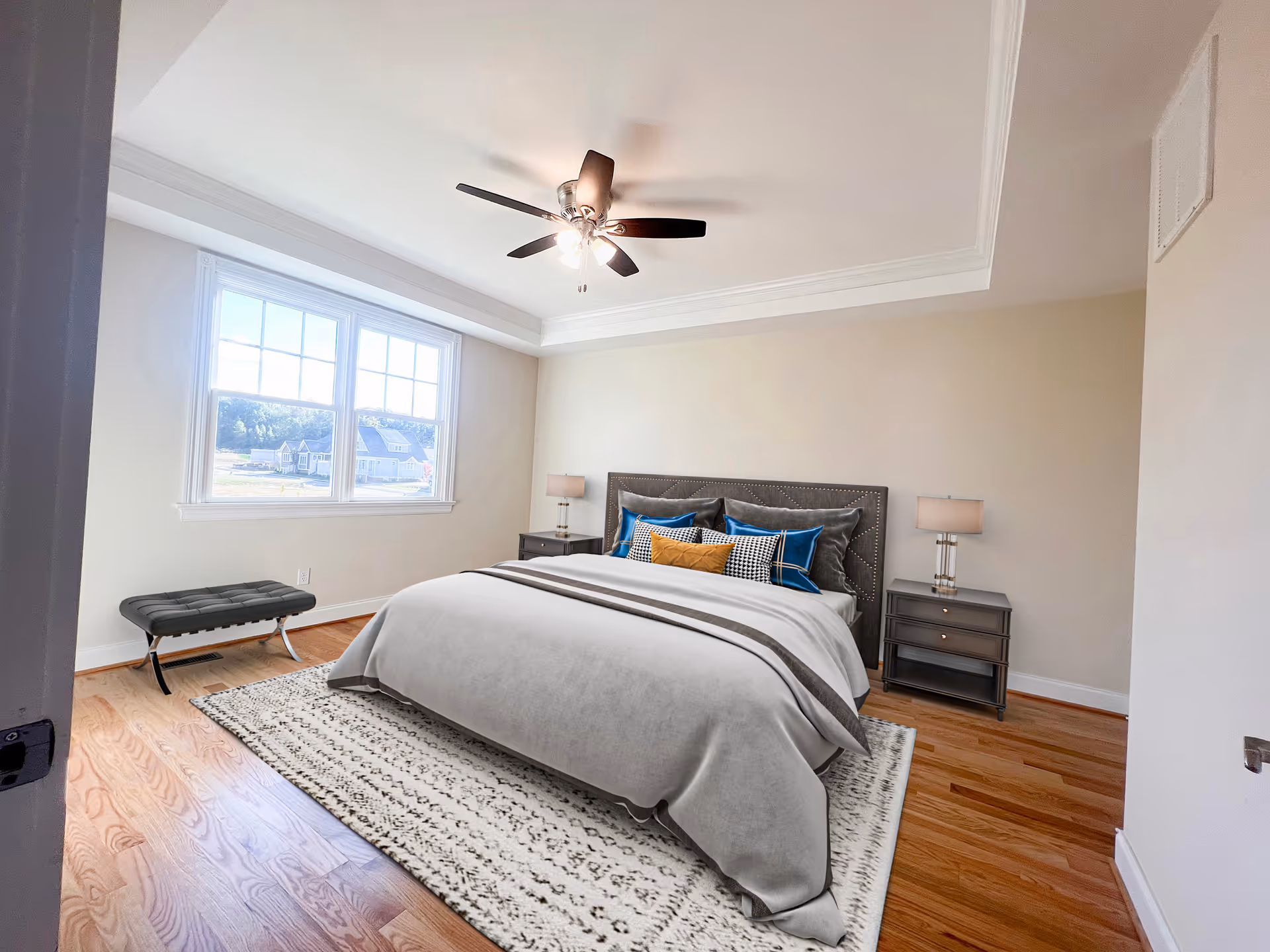 Bright bedroom with hardwood floors featuring a large bed with decorative pillows, two nightstands with lamps, a ceiling fan, and a window showing houses outside.
