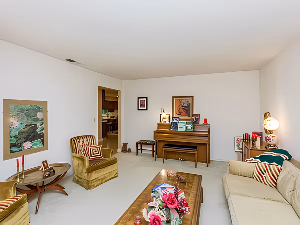 A living room with beige carpet and white walls featuring a beige sofa with striped pillows, two vintage armchairs with striped cushions, a wooden coffee table with a floral centerpiece, a wooden piano with framed pictures and a lamp on top, and a small wooden side table with a lamp and decorative items. There is a doorway leading to a kitchen area in the background.