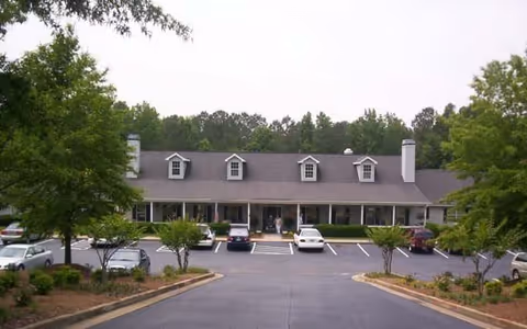 A long one-story building with dormer windows and a covered front porch facing a tree-lined parking lot with several parked cars.