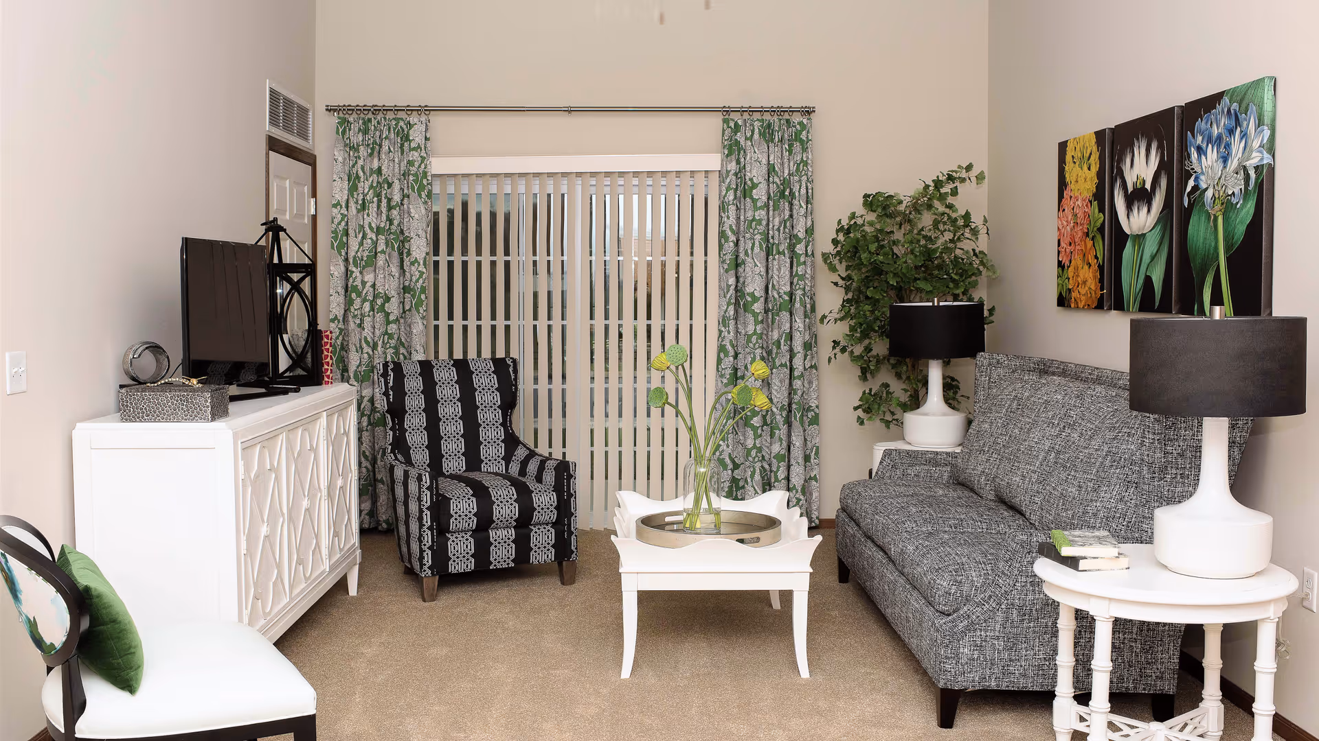 A cozy living room with a gray patterned sofa, a black and white patterned armchair, a white coffee table with a glass vase holding green and yellow flowers, and a white side table with a lamp and books. There is a white cabinet with a TV on top, green floral curtains framing a sliding glass door, and three colorful floral paintings on the wall. A large green plant is placed in the corner.