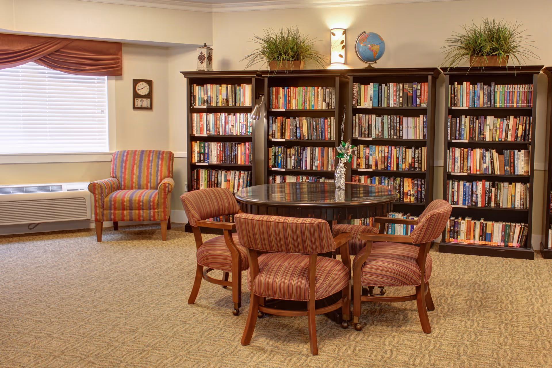 A cozy reading room with a round wooden table surrounded by four striped upholstered chairs. Behind the table are three tall dark wood bookshelves filled with books, decorated with two potted plants and a globe on top. To the left, there is a striped armchair near a window with closed blinds and a valance. The room has beige carpet and walls with soft lighting.