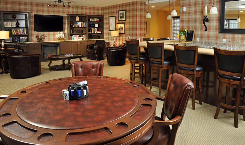 A cozy senior living common area featuring a round wooden game table with poker chips and cards, leather chairs, a bar with high stools, and a seating area with armchairs and bookshelves. The walls have a plaid pattern, and the room is warmly lit with lamps and pendant lights.