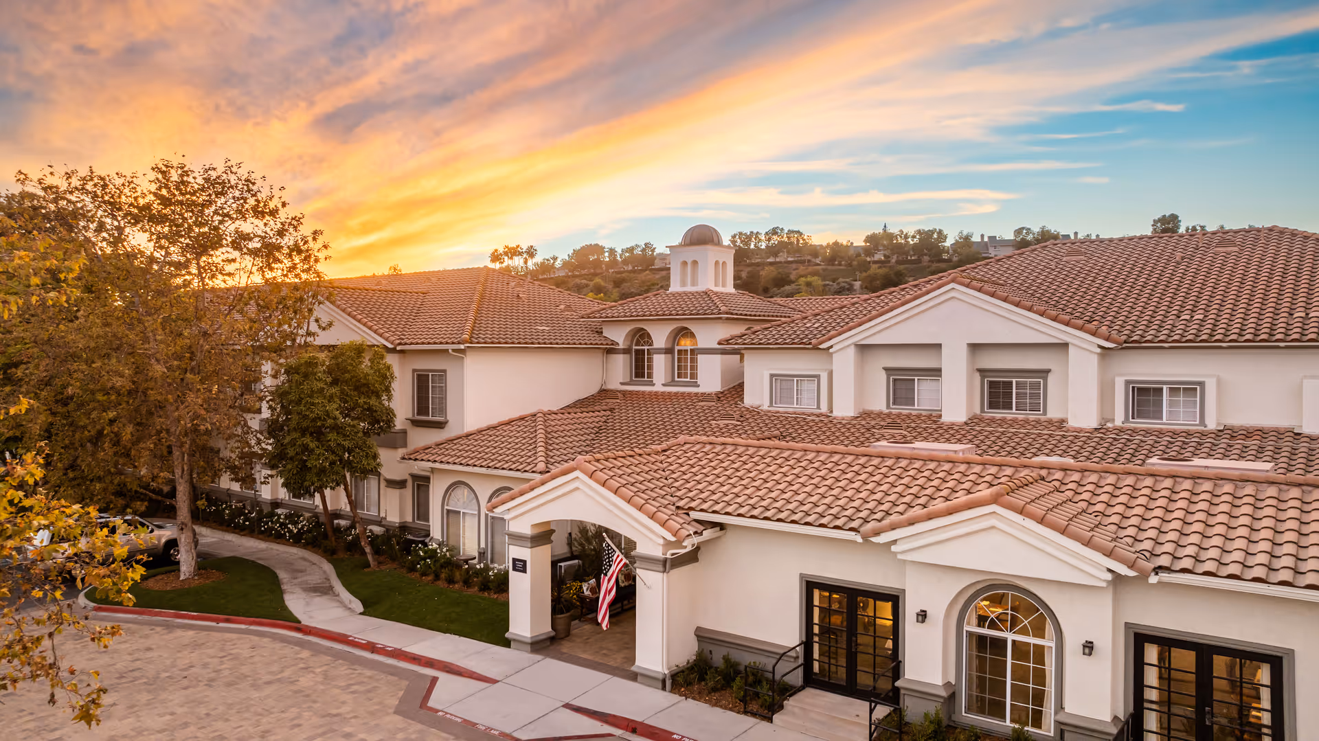 Exterior view of a senior living facility building with a tiled roof and arched windows during sunset, surrounded by trees and a curved driveway with an American flag near the entrance.