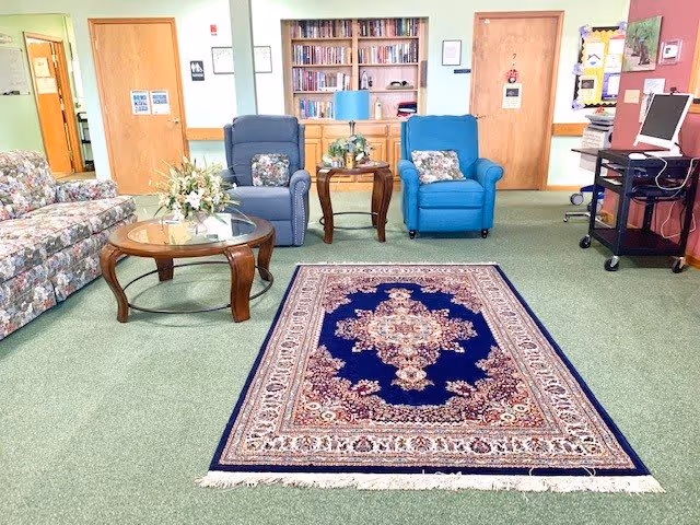 A cozy common area in a senior living facility featuring a floral-patterned sofa, two cushioned armchairs with decorative pillows, a glass-top wooden coffee table with a flower arrangement, a patterned area rug on green carpet, a bookshelf filled with books, and a computer workstation on a rolling cart against the wall.