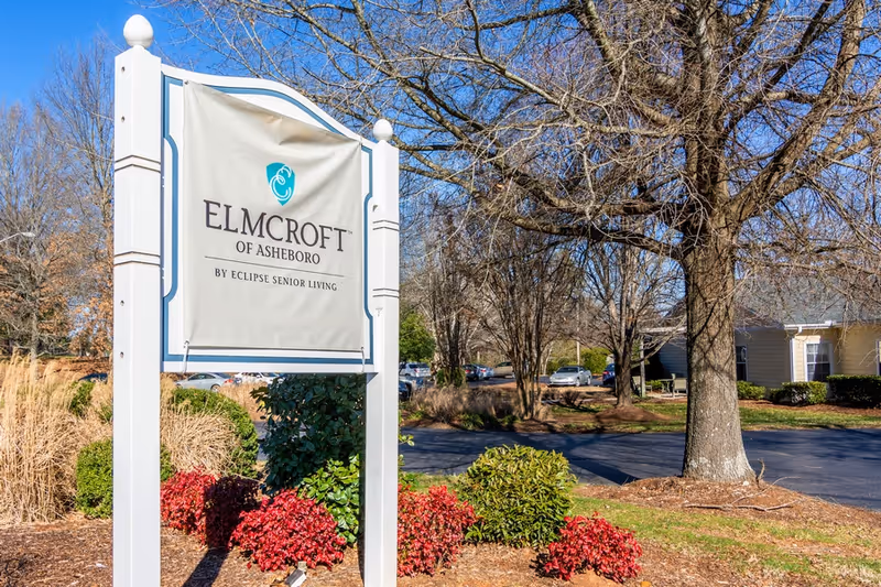Outdoor view of a white sign for Elmcroft of Asheboro by Eclipse Senior Living, surrounded by landscaping with bushes and trees, with a parking lot and buildings in the background under a clear blue sky.