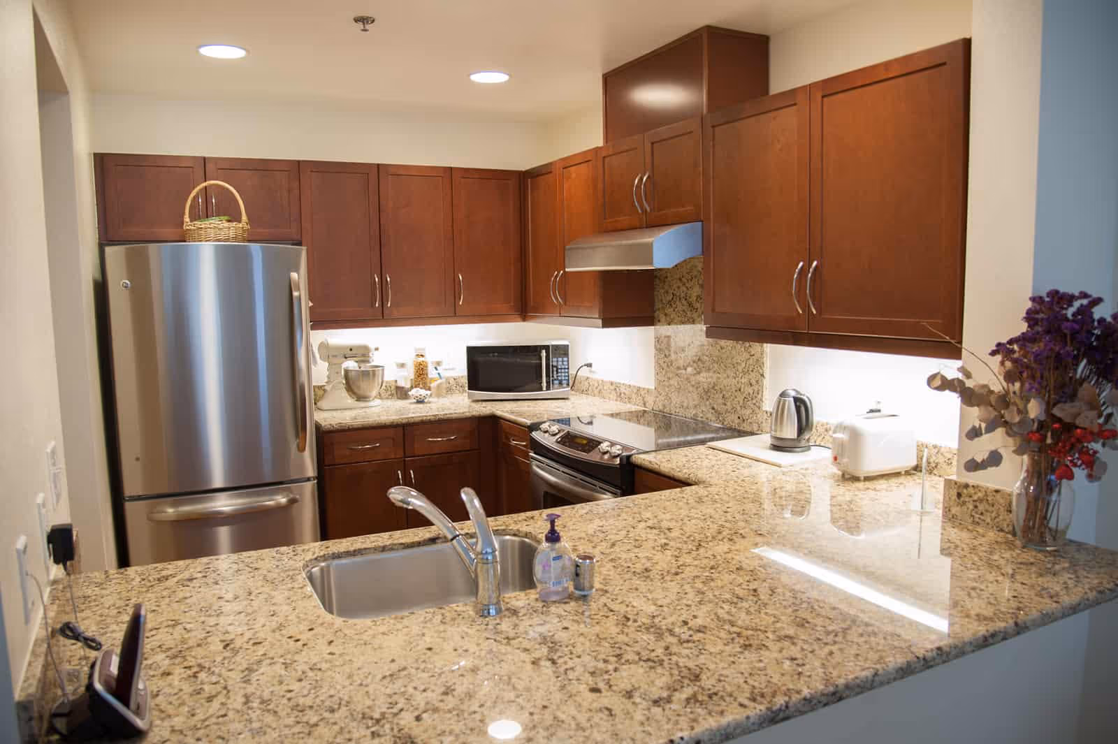 A modern kitchen with granite countertops, wooden cabinets, a stainless steel refrigerator, an electric stove with a range hood, a microwave, a mixer, a kettle, a toaster, and a vase with dried flowers on the counter.