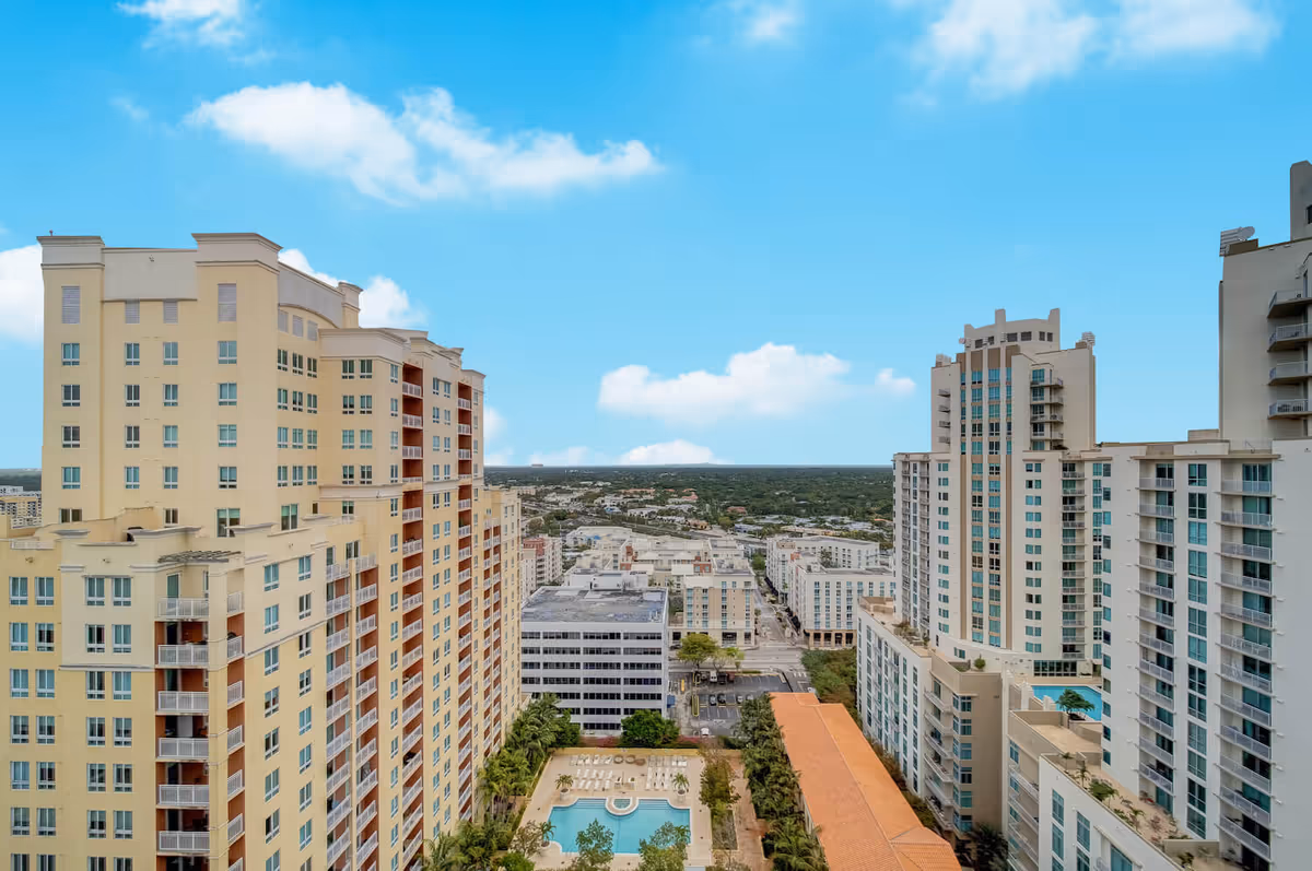 Aerial view of tall residential buildings around a courtyard pool with a cityscape and blue sky beyond.