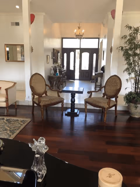 A cozy interior common area with two wooden armchairs with cushioned seats facing a small wooden table. The room has dark wooden flooring, a chandelier hanging from the ceiling, and a large front door with glass panels letting in natural light. There is a potted plant on the right side and a decorative mirror on the left wall.